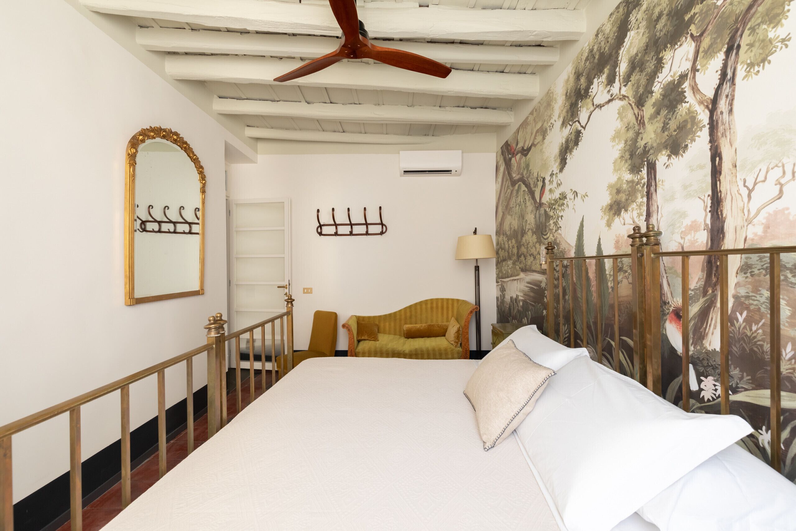 A bedroom with a large bed, a ceiling fan, a wall-mounted mirror, a chair, and a painting of trees.