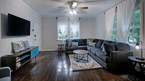 A living room with a sectional sofa, a flat-screen TV, a ceiling fan, and large windows with white curtains. - Walk to Downtown Fondren | King Beds (Jackson)
