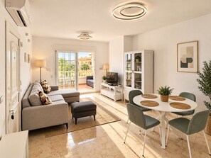 A modern living room with a sofa, ottoman, and dining table with chairs. There is a television on a stand, a cabinet, and a balcony with outdoor furniture. - Wonderful apartment with solarium terrace and community pools, Las Ramblas Golf (Orihuela Costa)