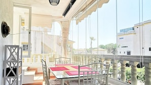 Un balcon avec une table et des chaises en verre, un luminaire suspendu et une vue sur des bâtiments et de la verdure.