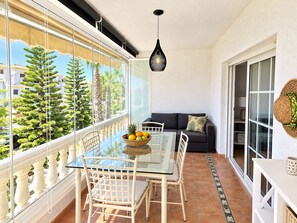 A balcony with a glass table, chairs, a basket of fruit, a hanging light fixture, and a view of trees and buildings. - Wonderful apartment with solarium terrace and community pools, Las Ramblas Golf (Orihuela Costa)