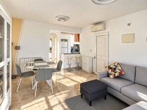 A modern living room with a grey sofa, a dining table with chairs, and a kitchen area with white cabinets and a stove.