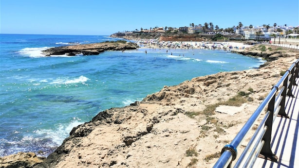 Une plage côtière au paysage rocheux, un ciel bleu dégagé et une vue sur une zone urbaine avec des bâtiments et des palmiers en arrière-plan.