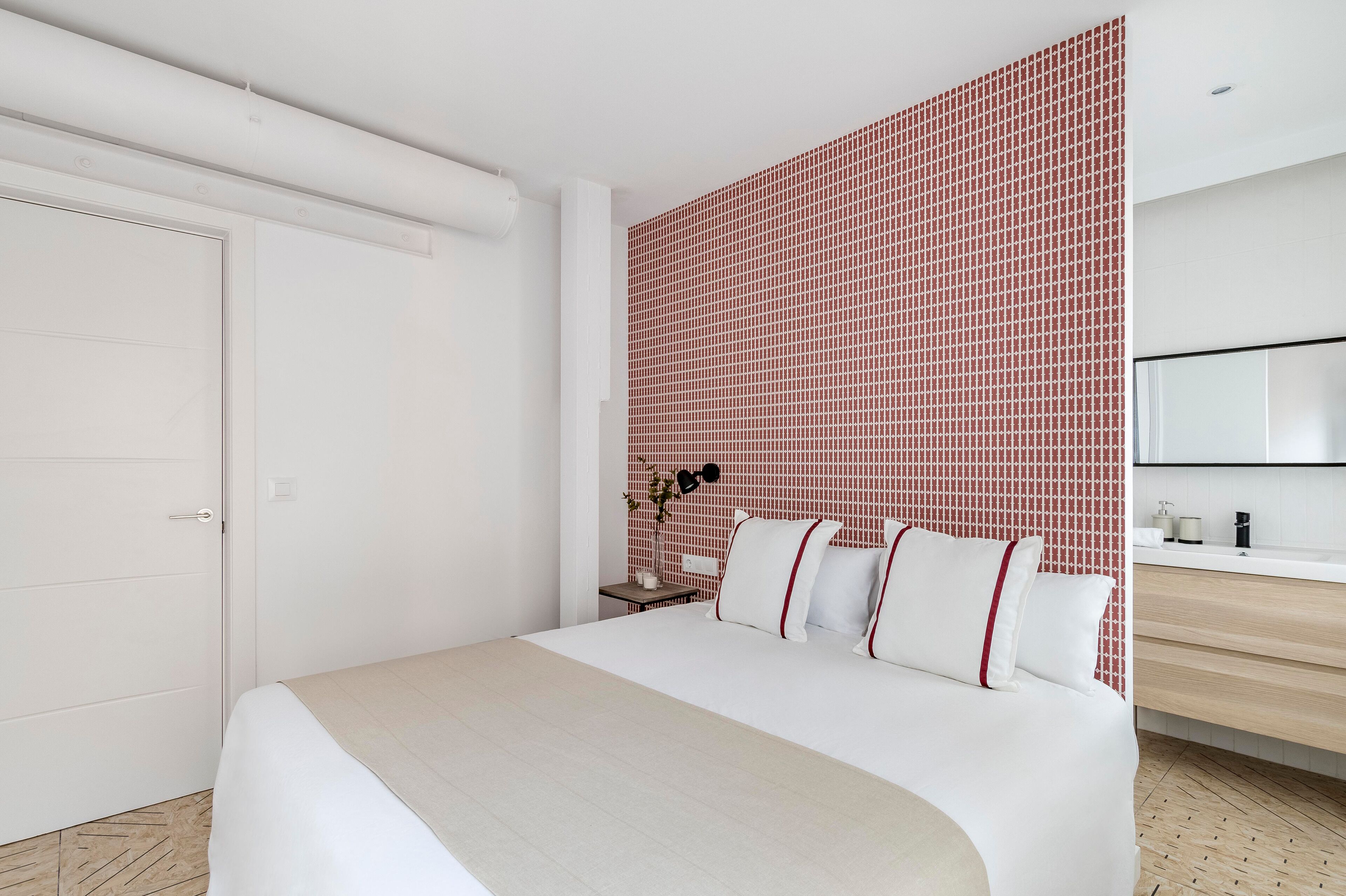 Una habitación de hotel moderna con una cama grande, una mesita de noche con lámpara, una pequeña planta y un baño con espejo y lavamanos.