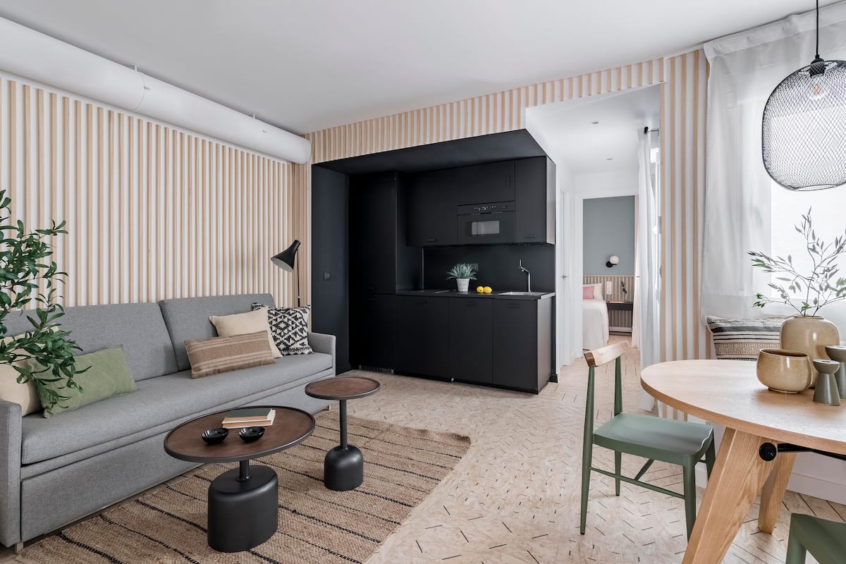 Uma sala de estar moderna com um sofá cinzento, uma mesa redonda de madeira e uma área de cozinha com armários pretos.
