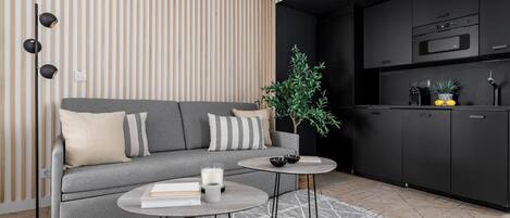 Een moderne woonkamer met een grijze bank, twee bijzettafeltjes, een potplant en een keuken met zwarte kasten en apparatuur.