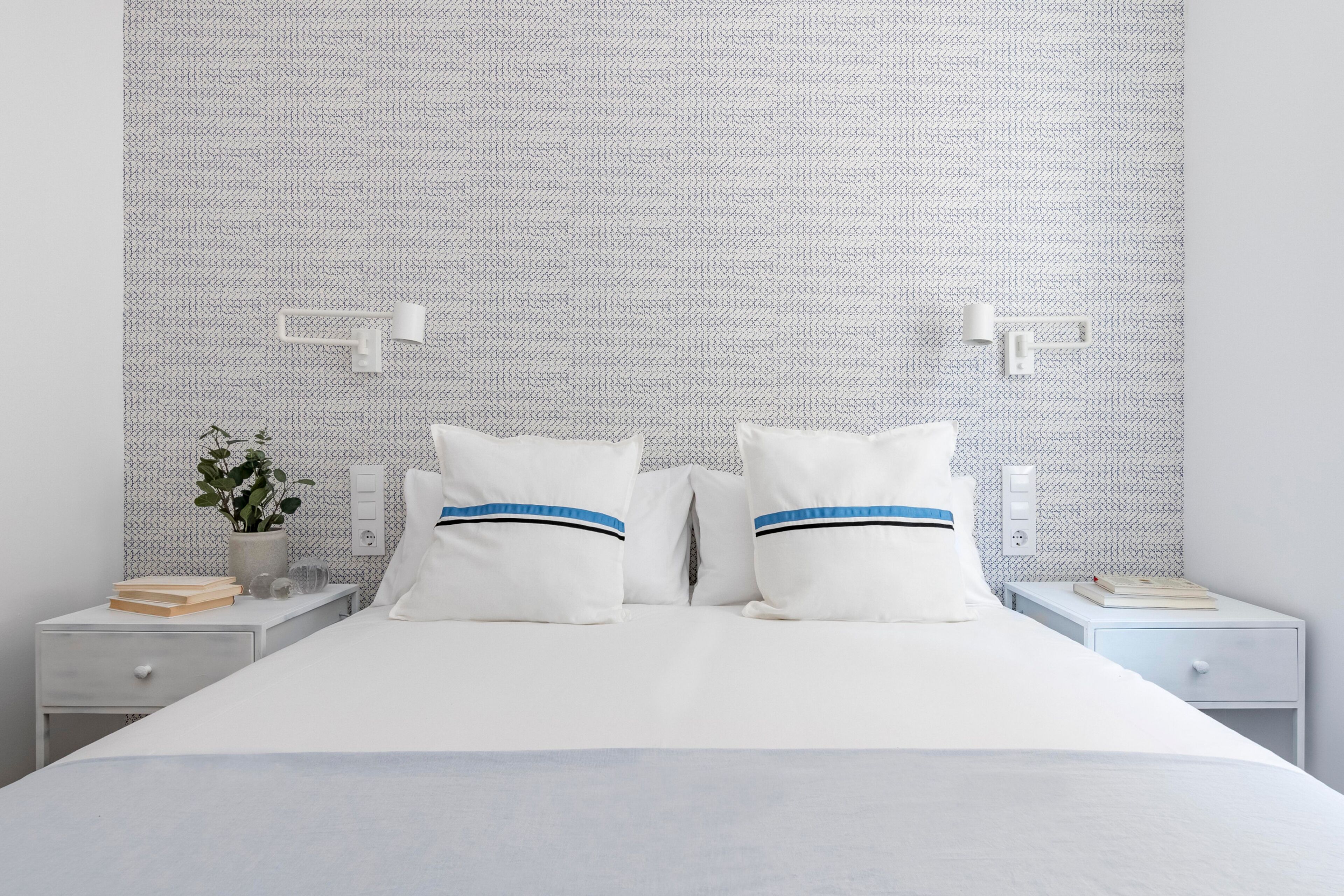 Una cama bien tendida con ropa blanca y dos almohadas, una mesita de noche con un libro y una planta, y una pared con un diseño texturizado.