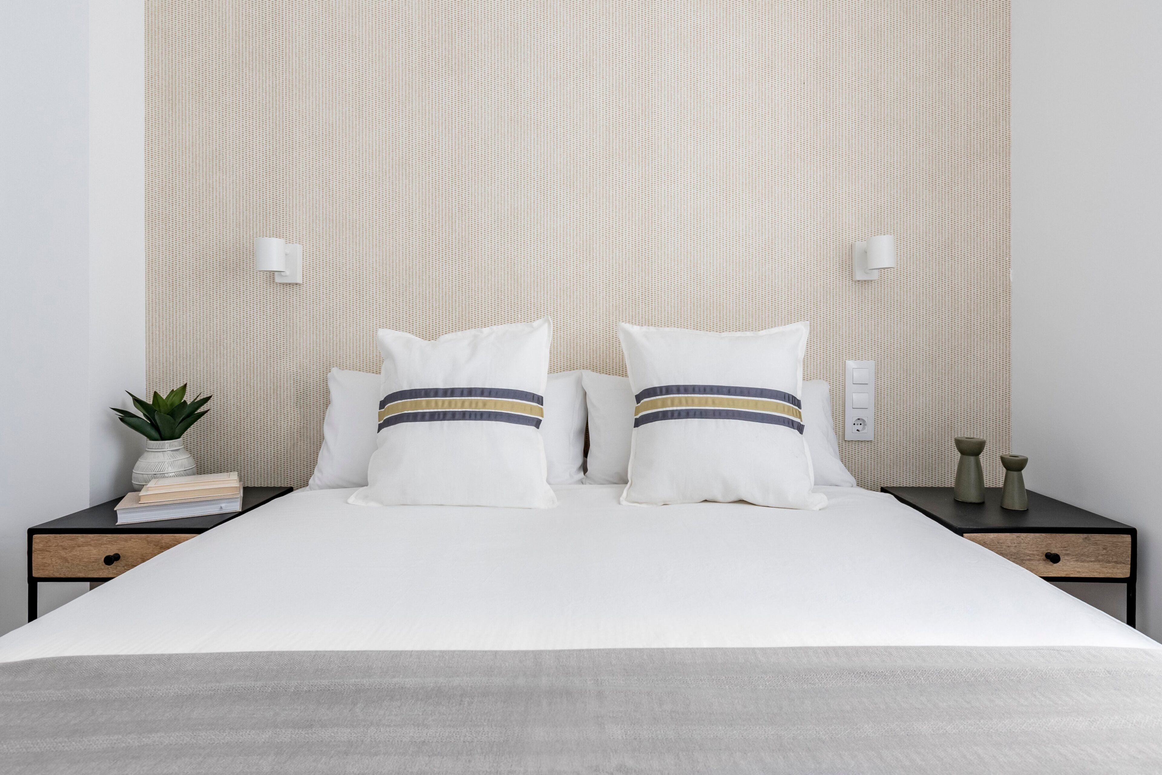 Una cama bien hecha con ropa blanca y dos almohadas de estampado a rayas. Hay una mesita de noche con una planta y un libro encima.
