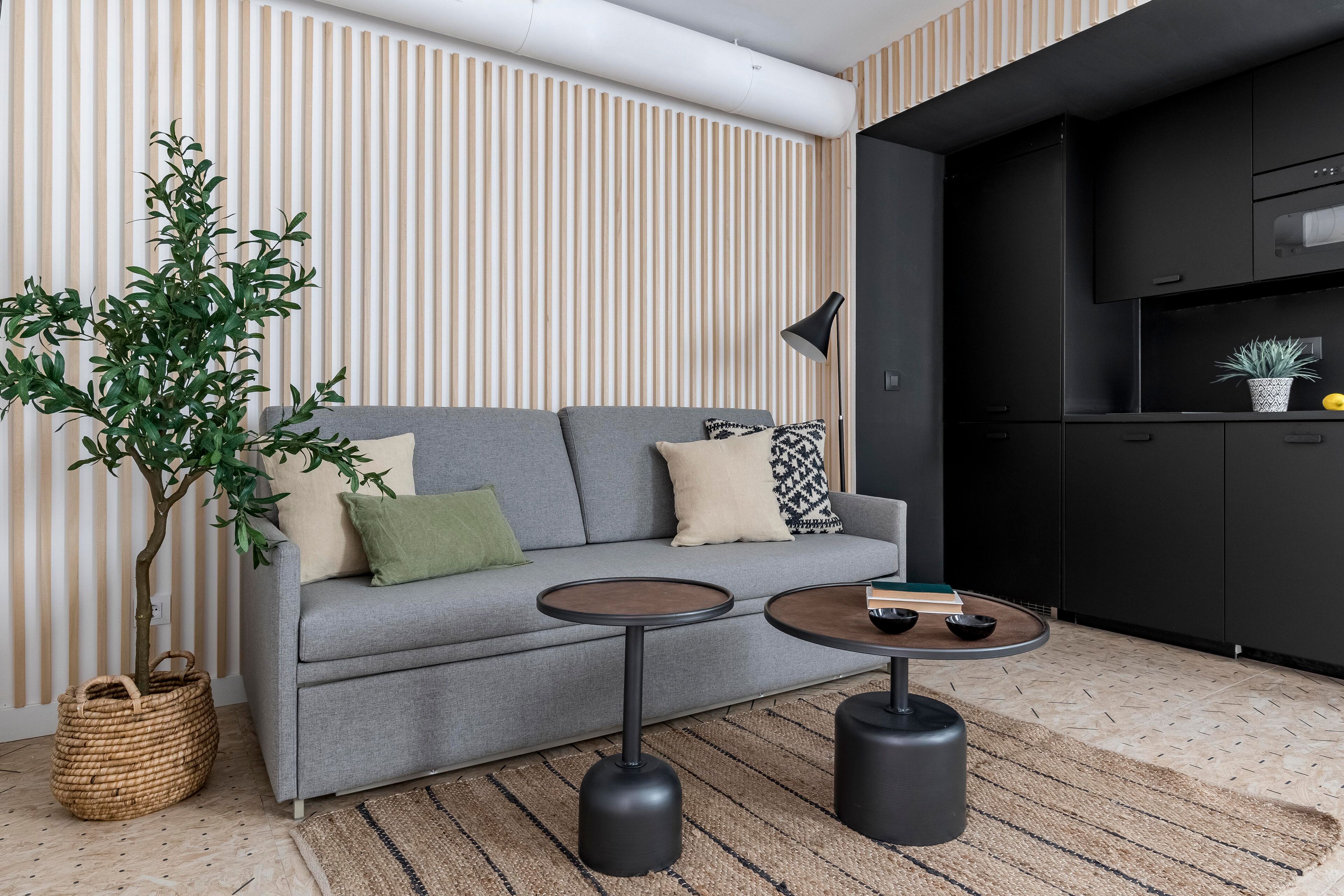 A modern living room with a grey sofa, two round tables, a potted plant, and a kitchen area with black cabinets.