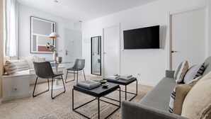 Ein modernes Wohnzimmer mit grauem Sofa, einem schwarzen Couchtisch, einem grauen Sessel und einem Esstisch mit Stühlen. Ein Flachbildfernseher ist an der Wand montiert und über der Tür hängt ein großer Spiegel.