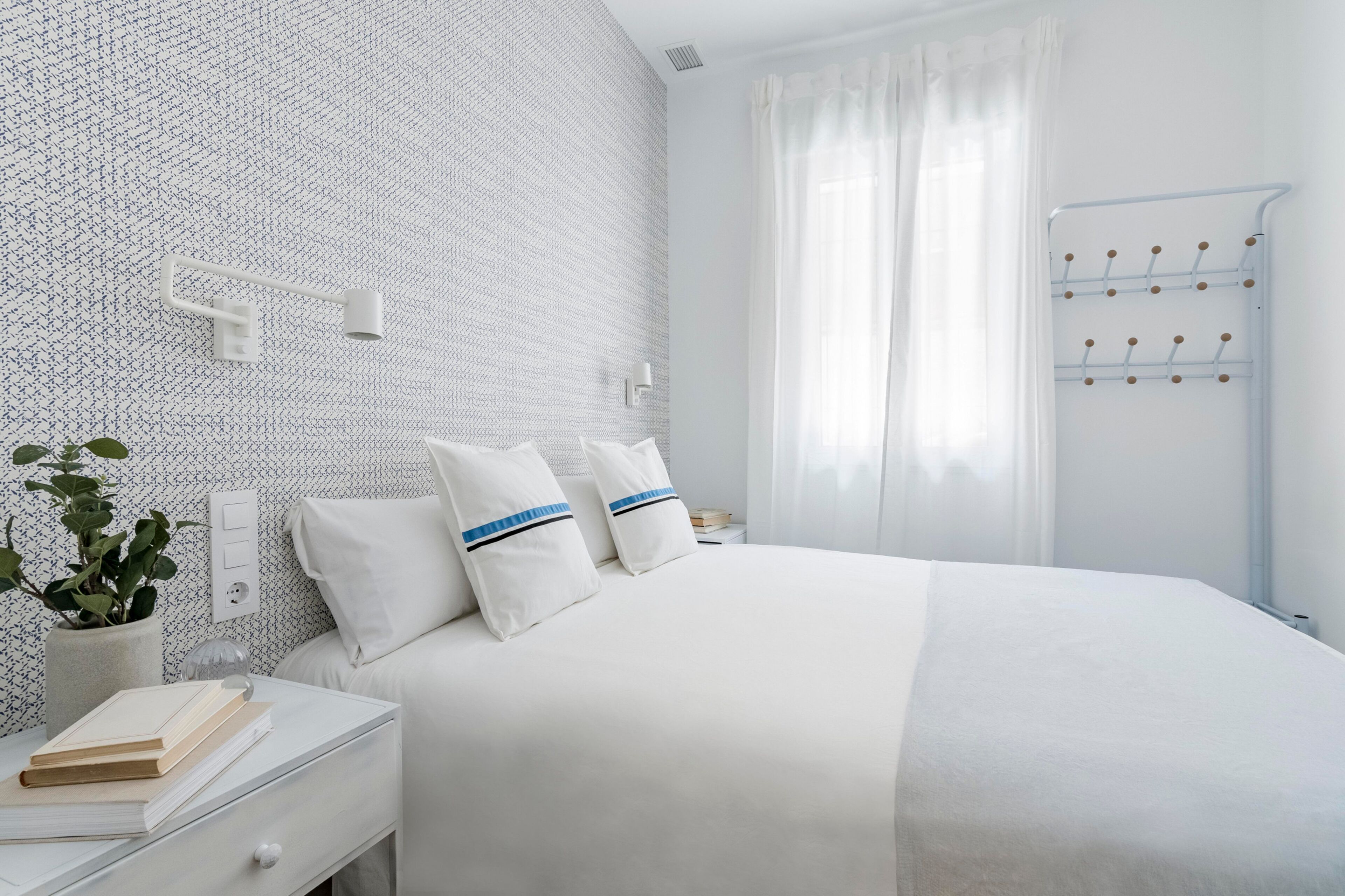 A neatly arranged bedroom with a patterned wall, a bedside table with books and a plant, and a window with curtains.