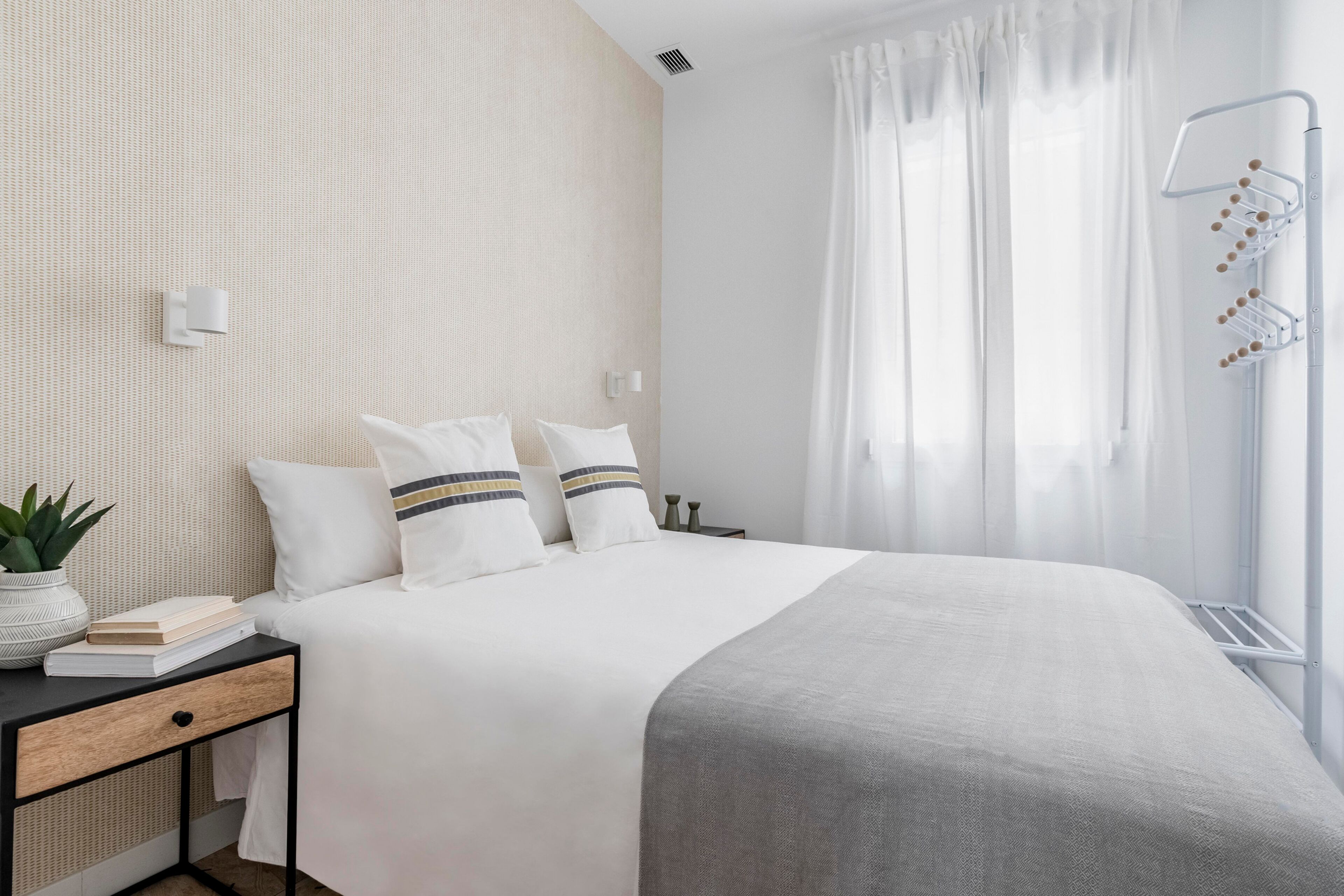 Een keurig opgemaakt bed met witte beddengoed en gestreepte kussens, een nachtkastje met een plant en boeken, en een raam met lichte gordijnen.