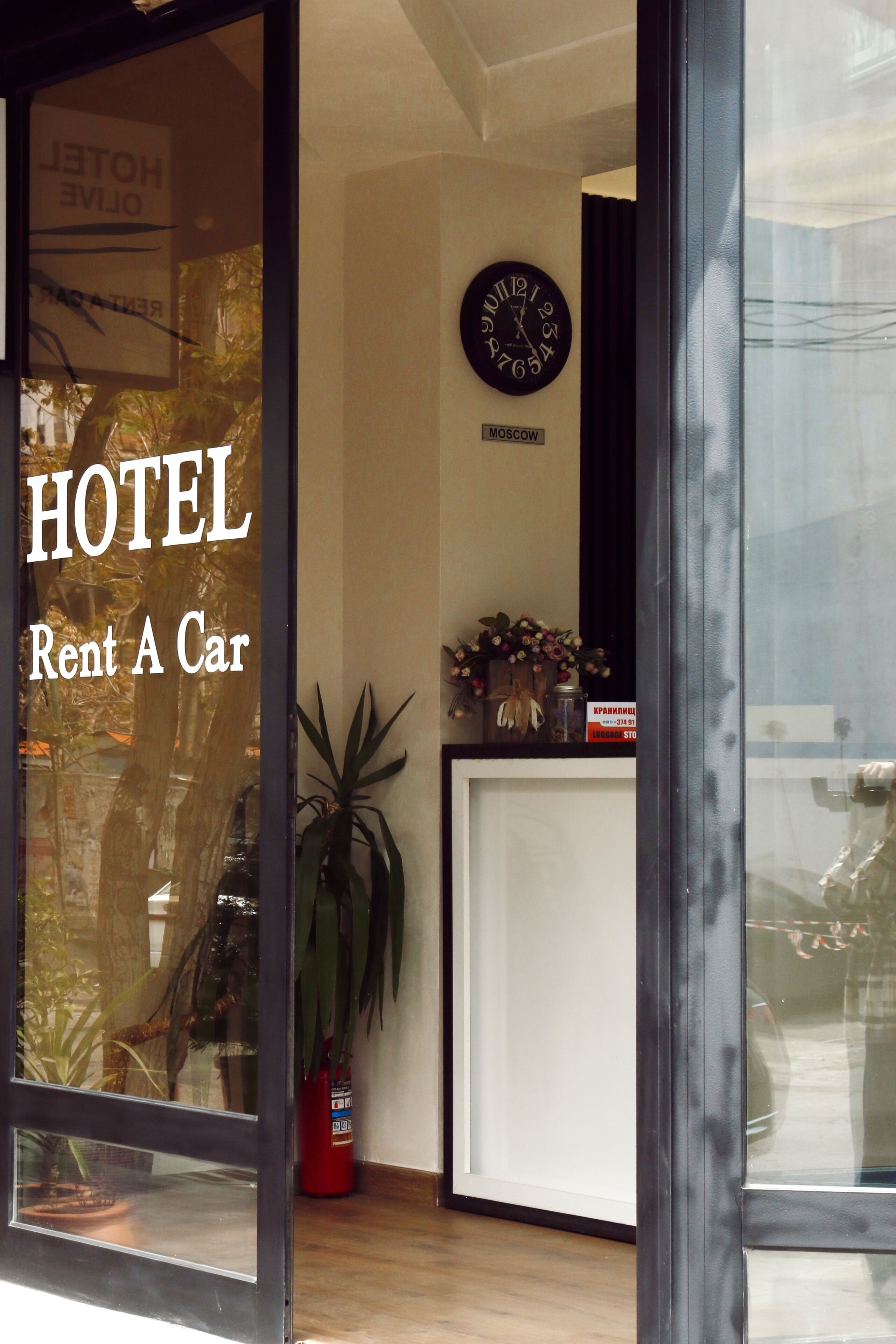 Een hotel receptie met een klok, een bord waarop 'HOTEL Rent A Car' staat, en een potplant.