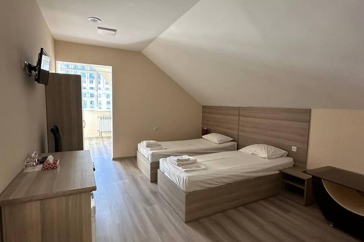 A modern, well-lit room with two beds, a desk, and a large window providing a view of buildings outside.