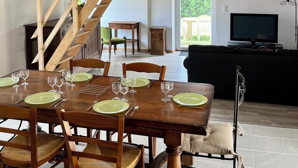 A dining area with a wooden table set for six, surrounded by wooden chairs. A staircase with a wooden railing leads to an upper level. - Maison du Périgord (La Douze)