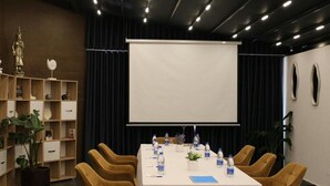 A conference room with a white table, yellow chairs, a projector screen, and a bookshelf with decorative items. - Hotel Alion21 (Durrës)