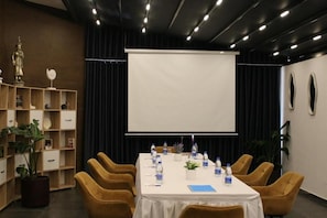 A conference room with a white table, yellow chairs, a projector screen, and a bookshelf with decorative items. - Hotel Alion21 (Durrës)