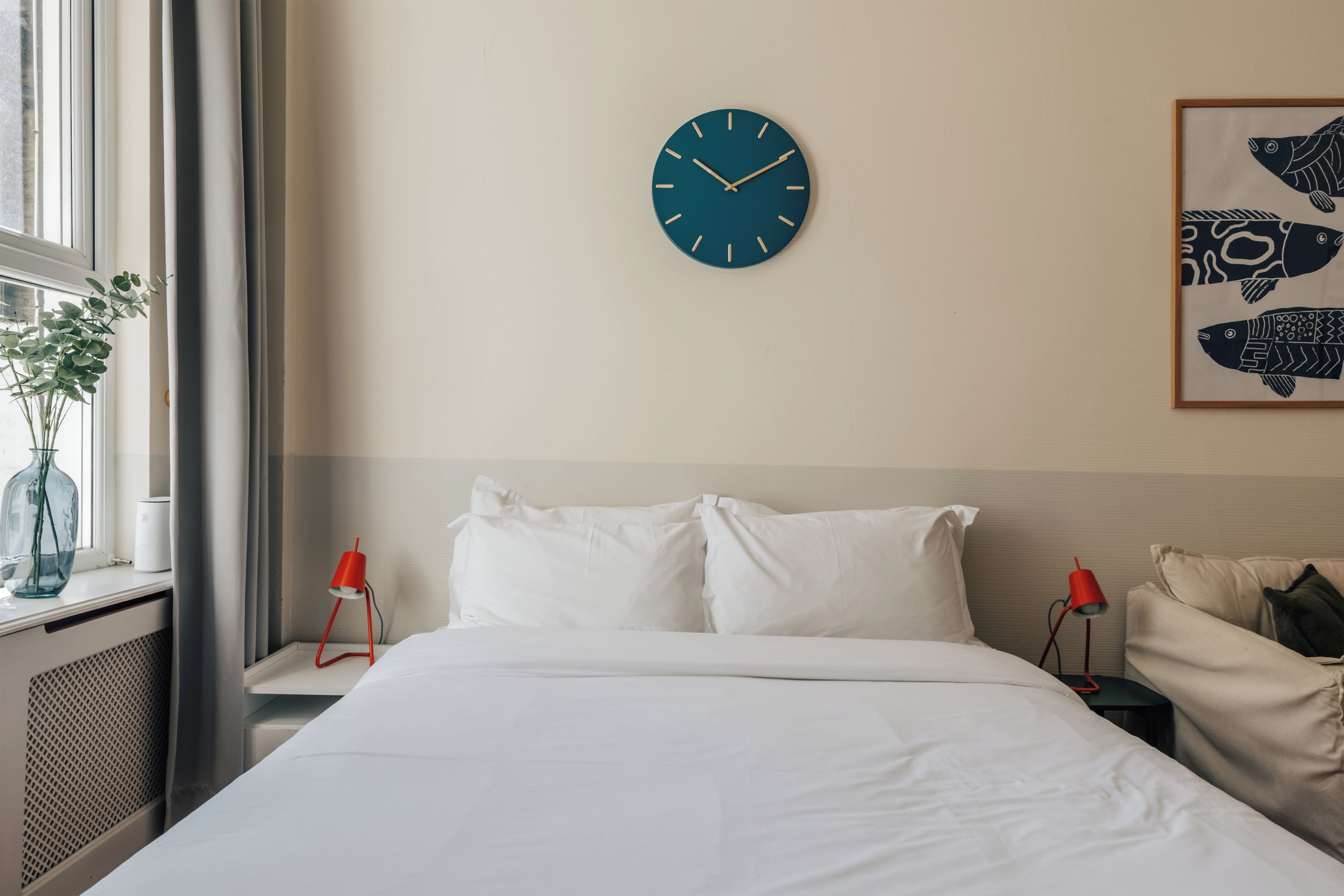 Camera d'albergo con un letto, una sveglia a parete, un quadro con soggetti ittici e una finestra con una pianta.