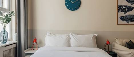 Camera d'albergo con un letto, una sveglia a parete, un quadro con soggetti ittici e una finestra con una pianta.