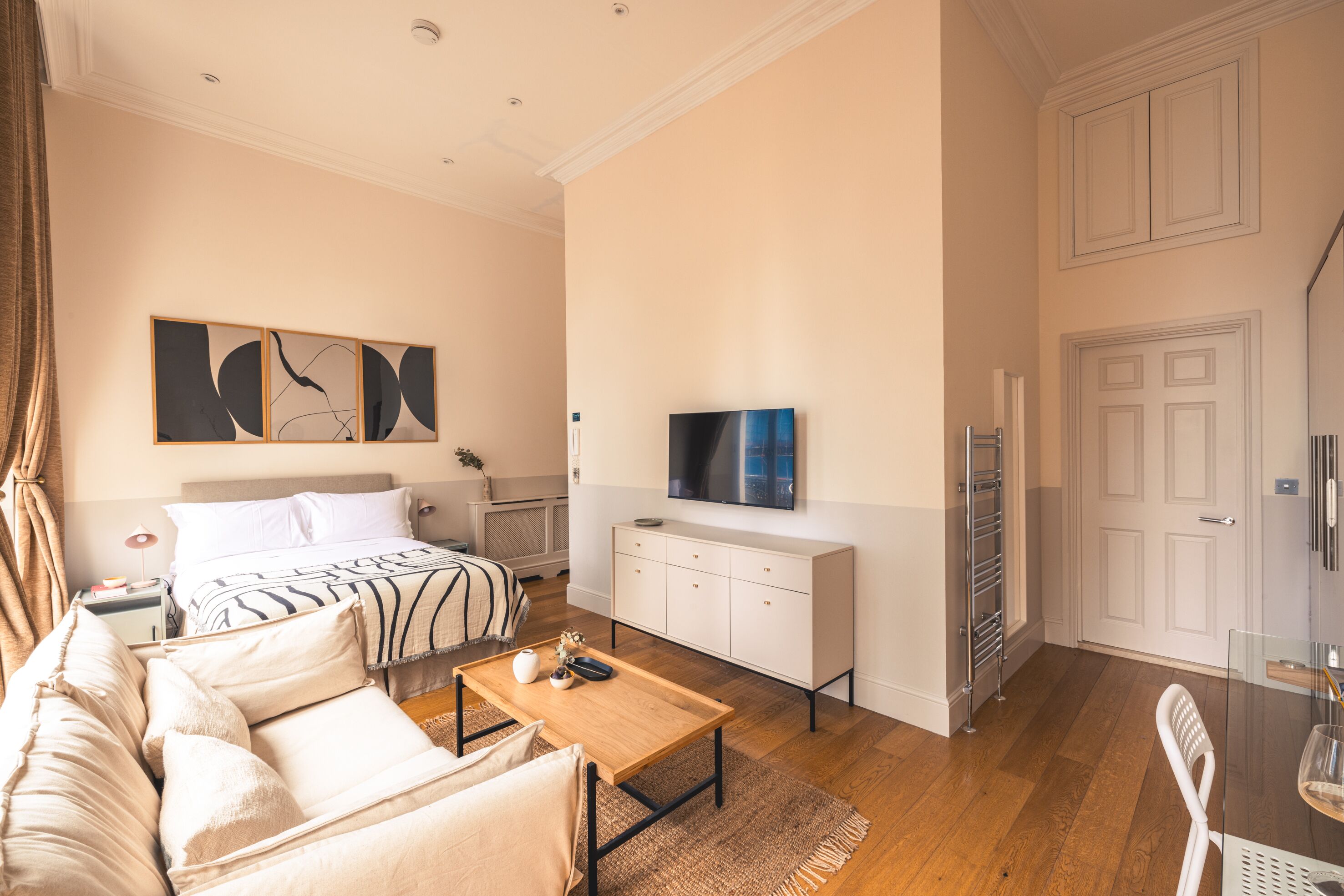 A modern hotel room with a bed, a sofa, a wooden coffee table, a TV, a white cabinet, and a door.