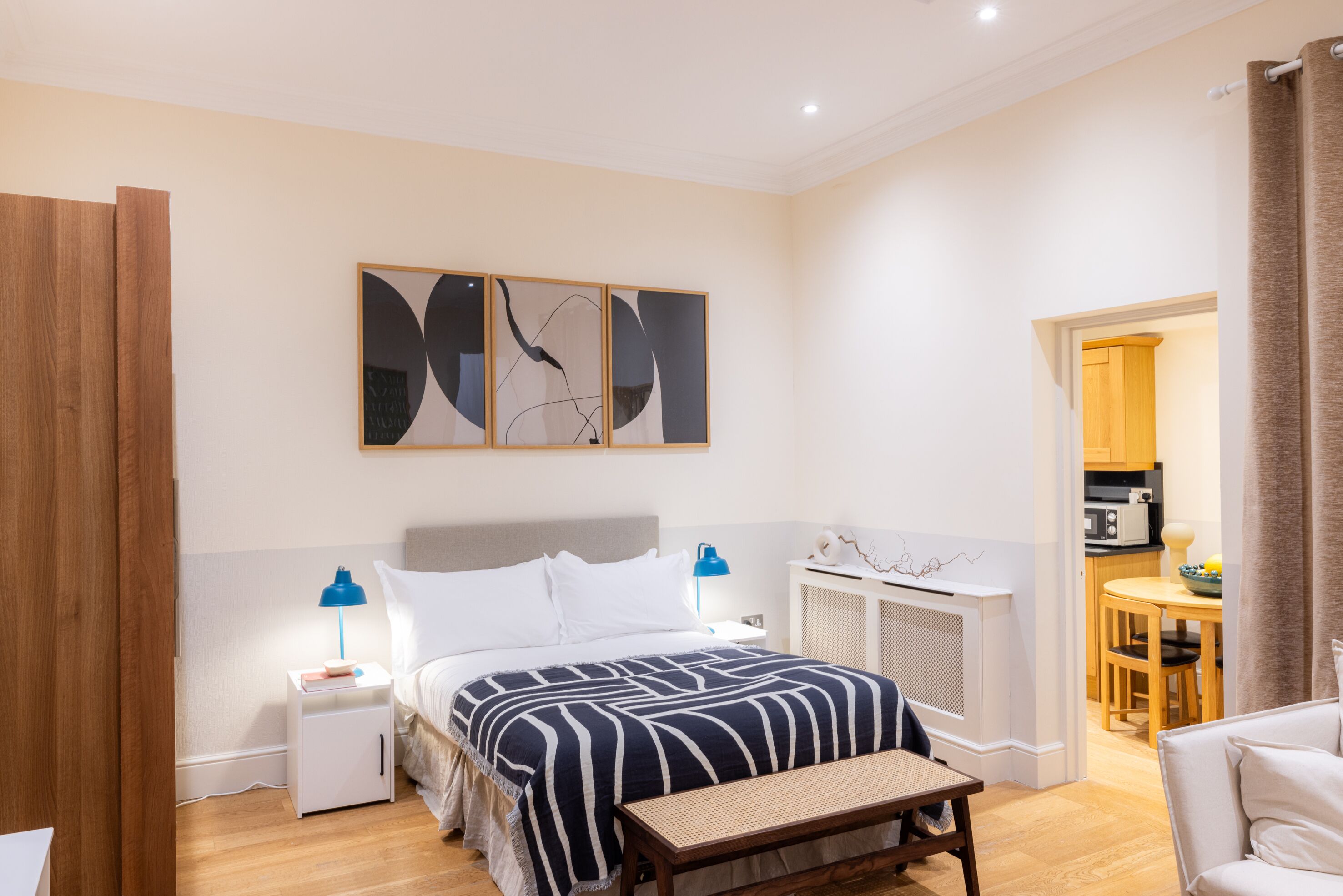 A bedroom with a bed, bedside tables, a bench, a wall-mounted artwork, and a view into a kitchen area.