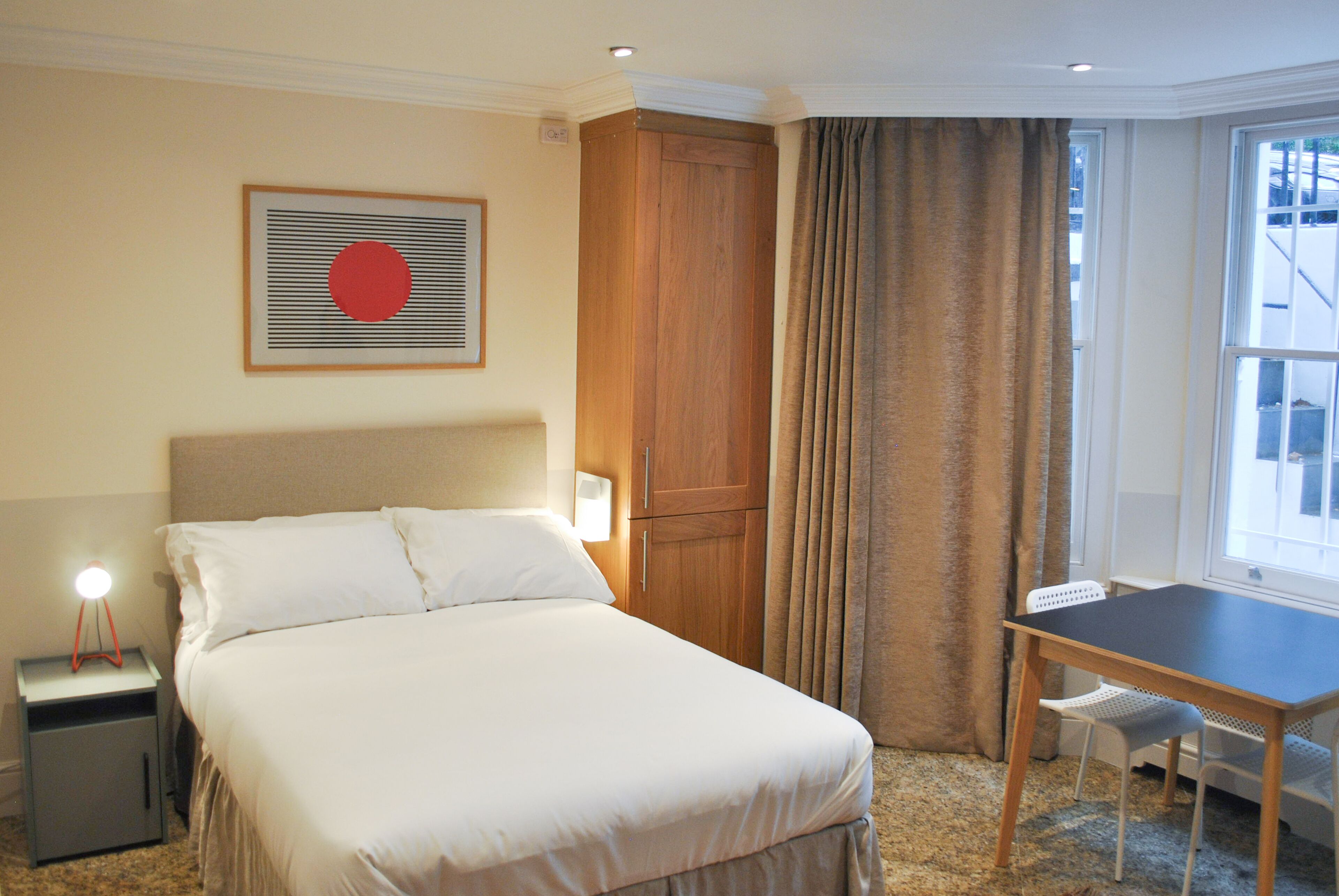 Habitación de hotel con cama, escritorio, silla, mesita de noche, lámpara, ventana con cortinas y un cuadro en la pared.