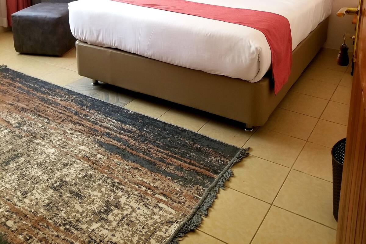 A neatly arranged hotel room with a large bed, a chair, a small ottoman, and a patterned rug.