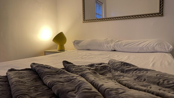 Una cama bien hecha con almohadas blancas y una manta gris, una mesita de noche con una lámpara amarilla y un espejo en la pared.