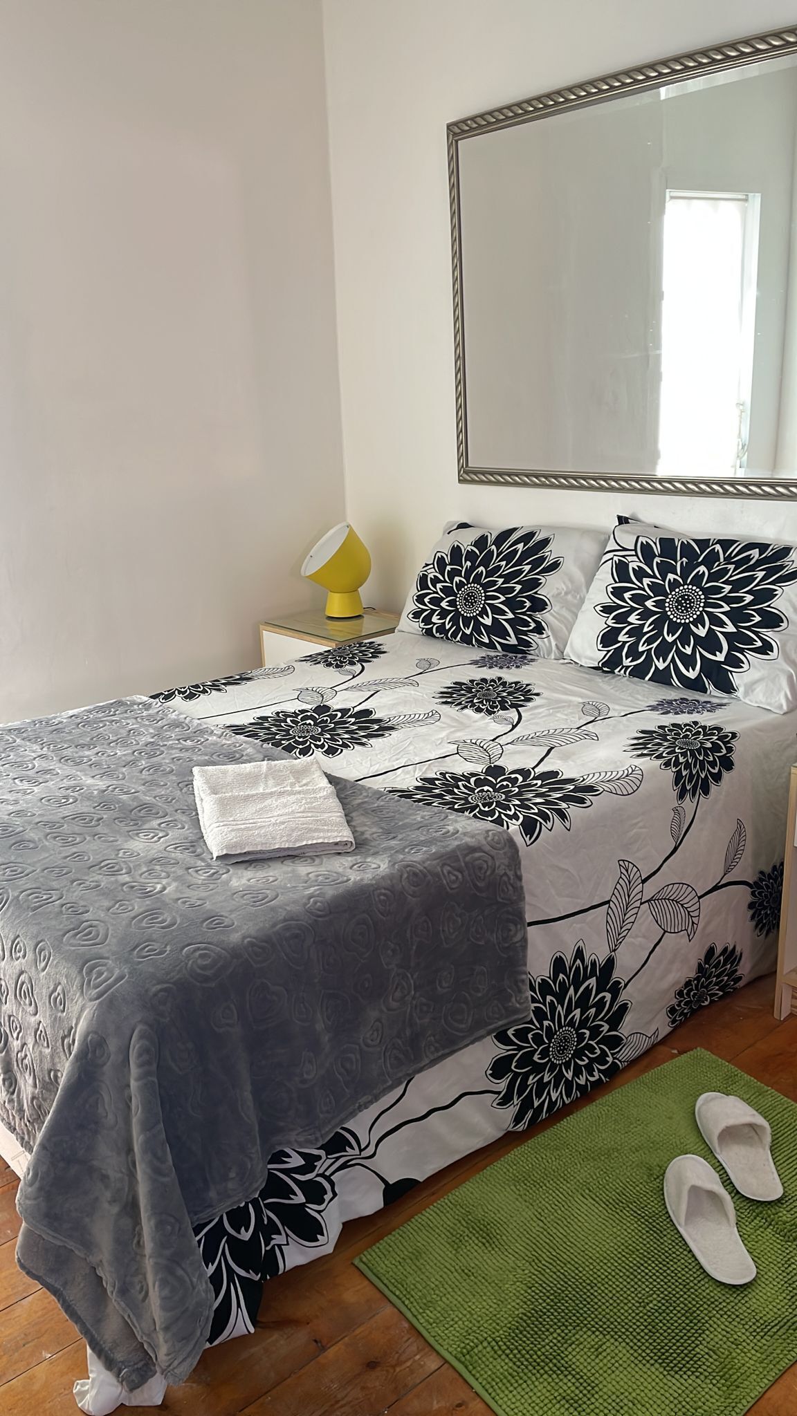 Ein ordentlich gemachtes Bett mit floralem Überwurf, einem grünen Teppich und einem Paar Hausschuhen auf dem Boden.