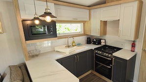 A compact kitchen with a microwave, stove, sink, and wooden cabinets. - Osprey Static Caravan - 2 Bedroom - No Pets (Prenteg)