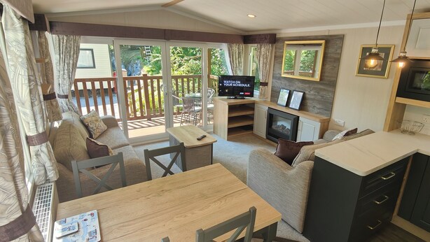 A cozy living area with a sofa, a wooden table, and a television. There is a fireplace and a balcony with a wooden railing. - Osprey Static Caravan - 2 Bedroom - No Pets (Prenteg)