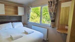 A bedroom with a bed, white linens, a wooden headboard, a window with curtains, and built-in storage cabinets. - Osprey Static Caravan - 2 Bedroom - No Pets (Prenteg)