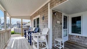 A covered porch with Adirondack chairs and a small table, wooden deck flooring, and a brick wall. - Welcome To Tidal Fix! Classic Sea Side Cottage With Modern Comforts RPAM094 (Harwich)