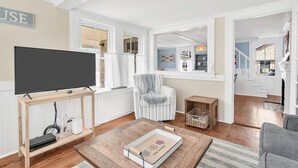 A living room with a TV, a striped armchair, a coffee table, and a view of the outside. - Welcome To Tidal Fix! Classic Sea Side Cottage With Modern Comforts RPAM094 (Harwich)