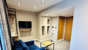 Ein modernes Wohnzimmer mit einem blauen Sofa, einem kleinen Tisch mit Pflanze, einem an der Wand befestigten Flachbildfernseher und einer Holztür im Hintergrund.