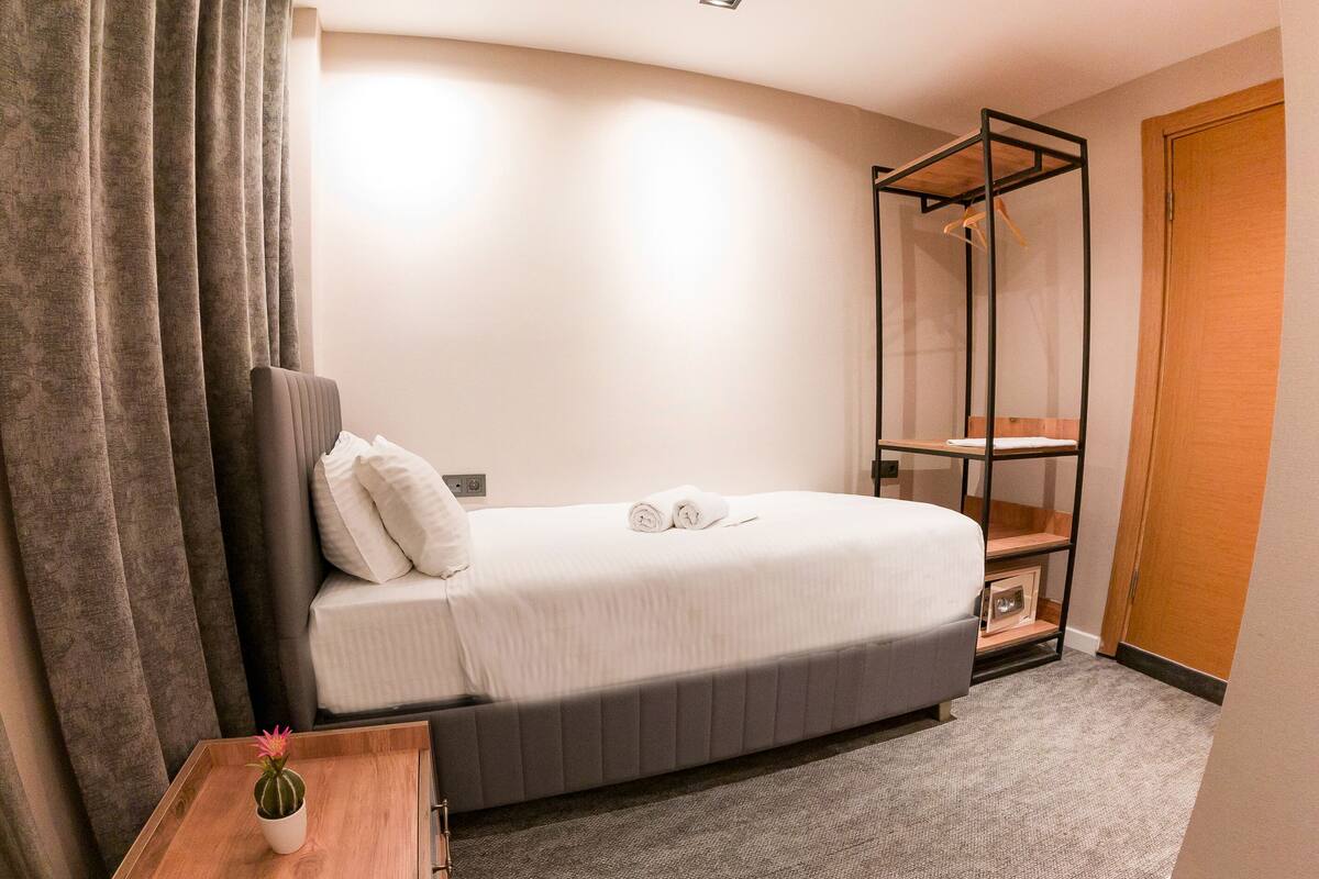 A neatly made bed with pillows and a rolled towel, a bedside table with a small potted plant, a wooden wardrobe, and a metal shelf unit.