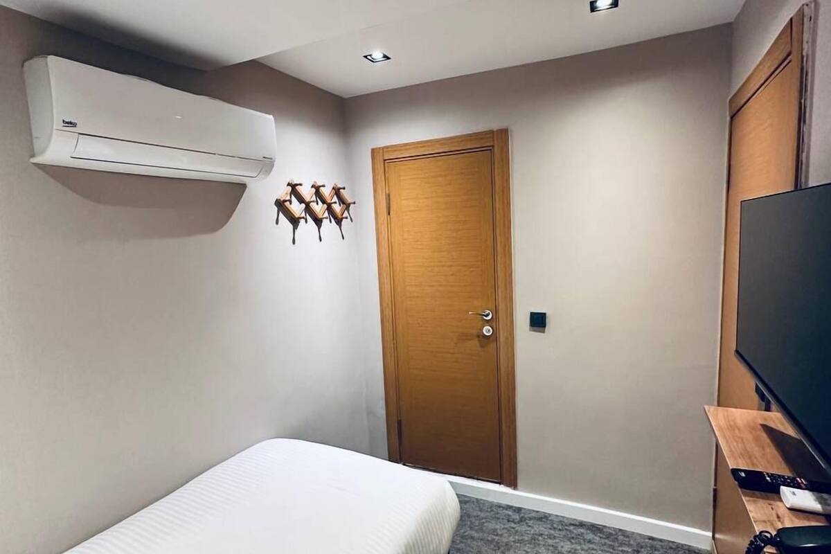 A hotel room with a bed, an air conditioning unit, a wooden door, a television on a wooden stand, and a carpeted floor.