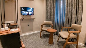 Ein Hotelzimmer mit Fernseher, der die Serie 'Friends' zeigt, einem Holznachttisch mit Wasserkocher und Tassen, zwei Holzstühlen und einem großen Fenster mit Vorhängen.