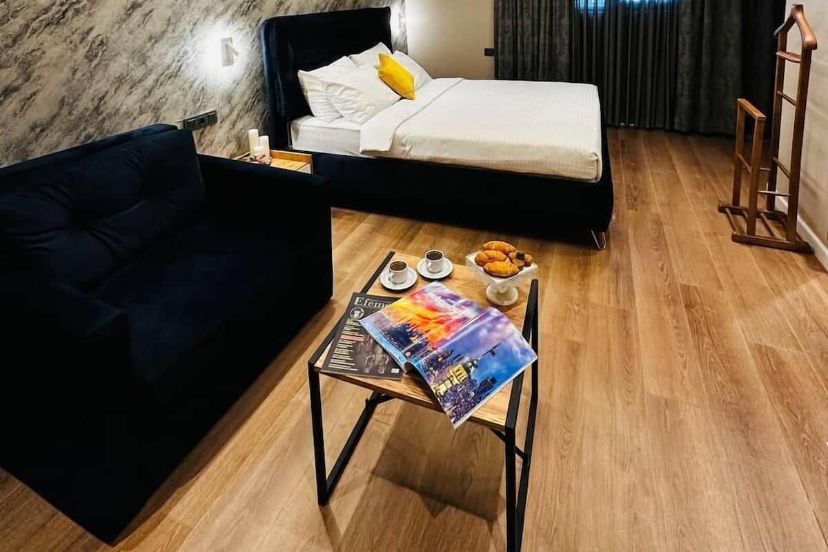 A modern hotel room with a bed, a dark-colored sofa, a small table with a magazine, and a wooden floor.