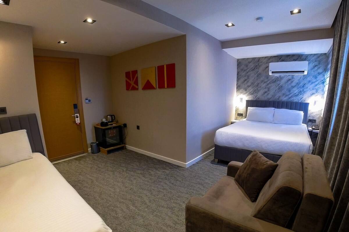 A hotel room with two beds, a sofa, a TV, and an air conditioning unit.