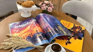 Ein Tisch mit einer Zeitschrift, einer Kaffeetasse, einem Teller mit Gebäck und einem Blumenstrauß.