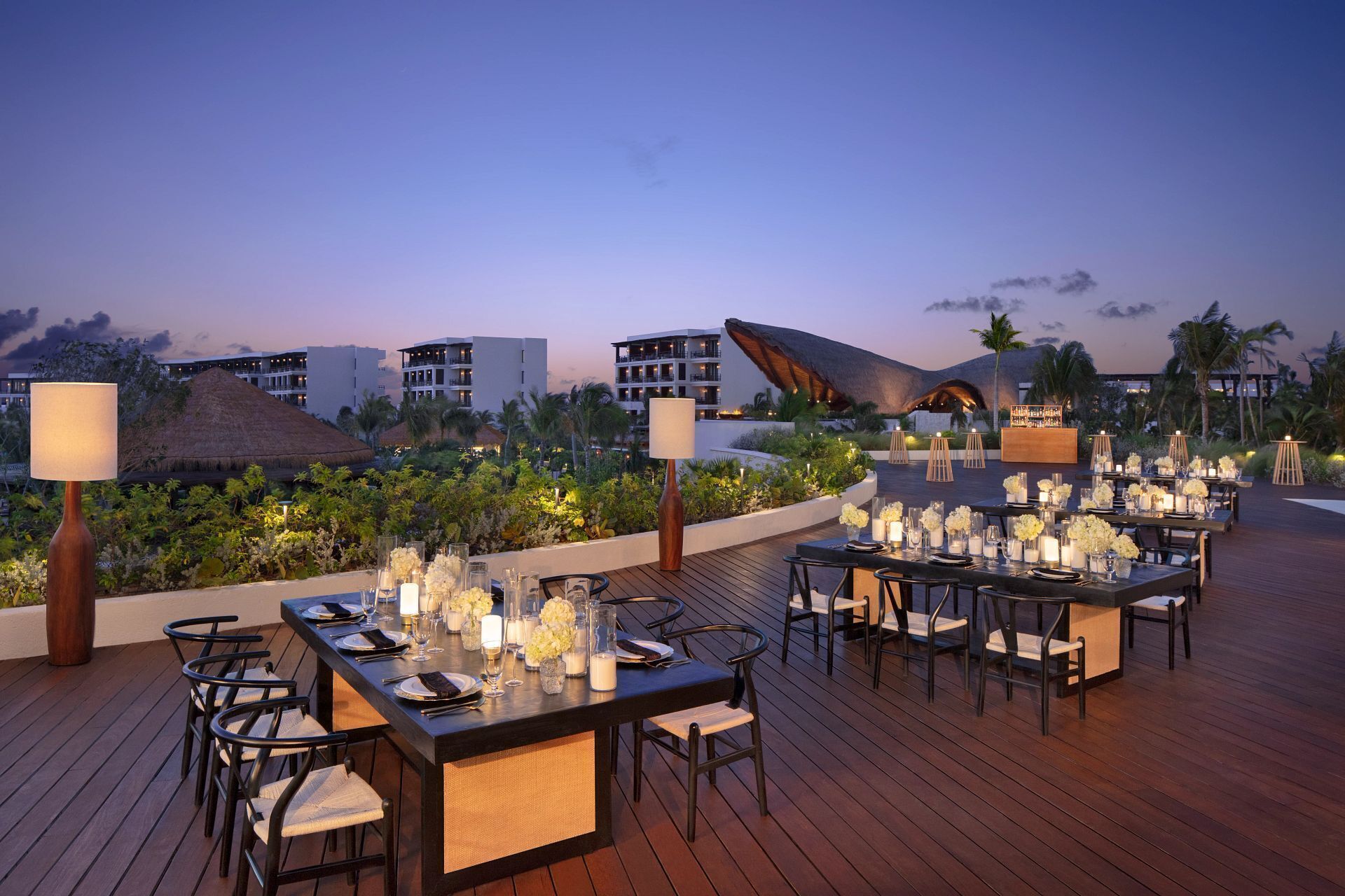 An outdoor dining area with tables set for a meal, surrounded by lush greenery and overlooking a resort with multiple buildings.