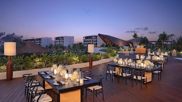 An outdoor dining area with tables set for a meal, surrounded by lush greenery and overlooking a resort with multiple buildings.