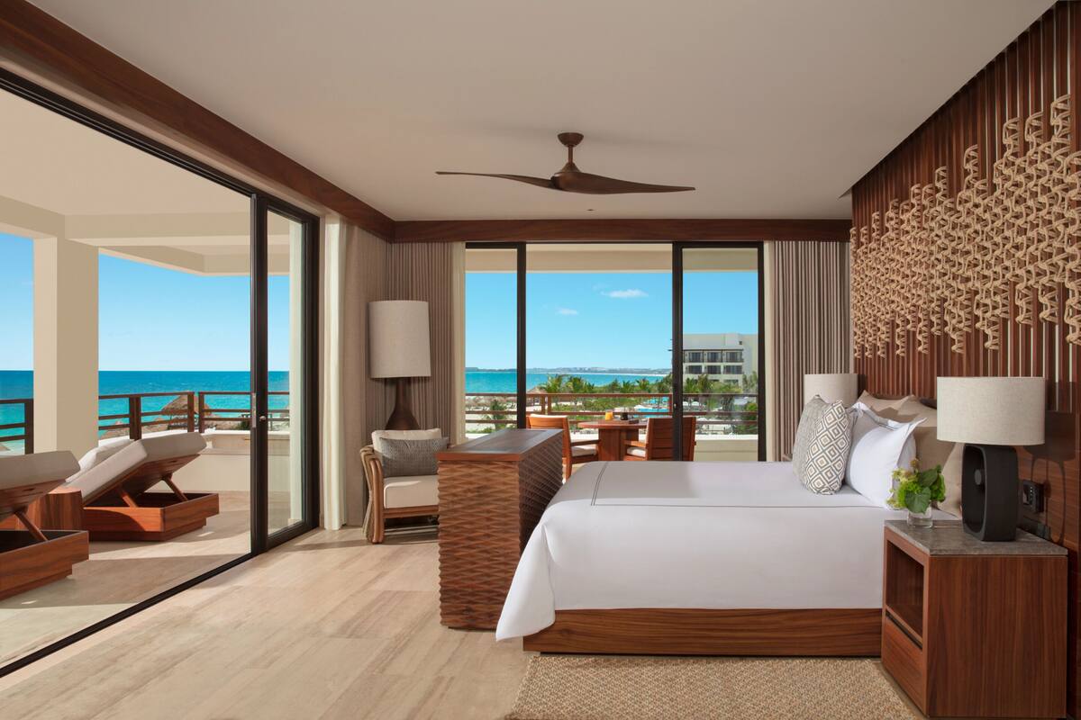 A hotel room with a large bed, a ceiling fan, a view of the ocean, and a balcony with lounge chairs.