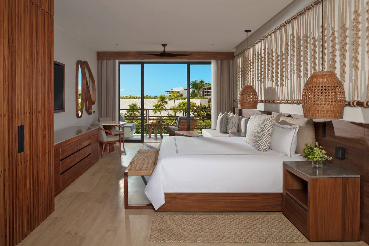A modern hotel room with a large bed, wooden furniture, a balcony with a view of palm trees and buildings, and a ceiling fan.