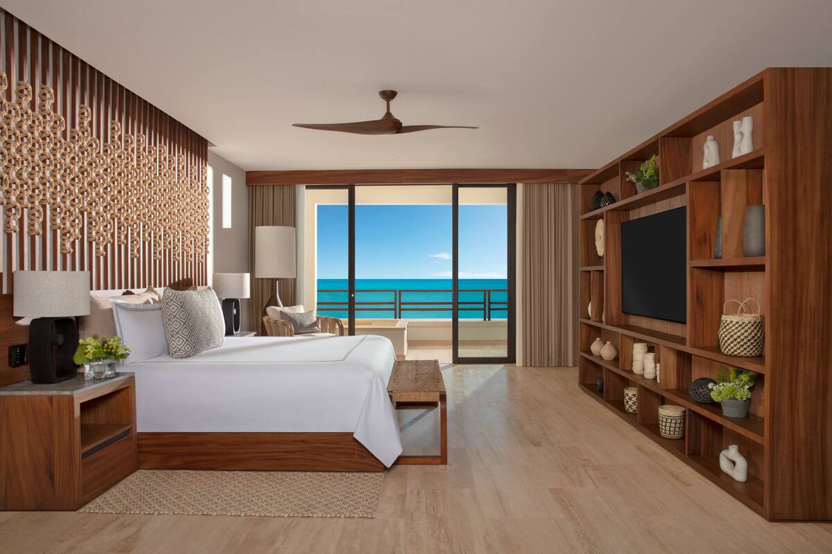 A modern hotel room with a large bed, a TV, wooden shelves, and a balcony with a view of the ocean.