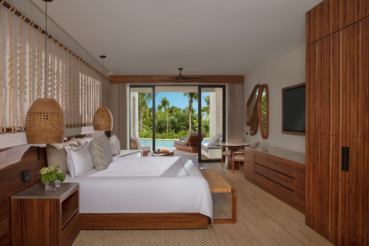 A spacious bedroom with a large bed, a wooden wardrobe, a television, and a view of a pool and palm trees.