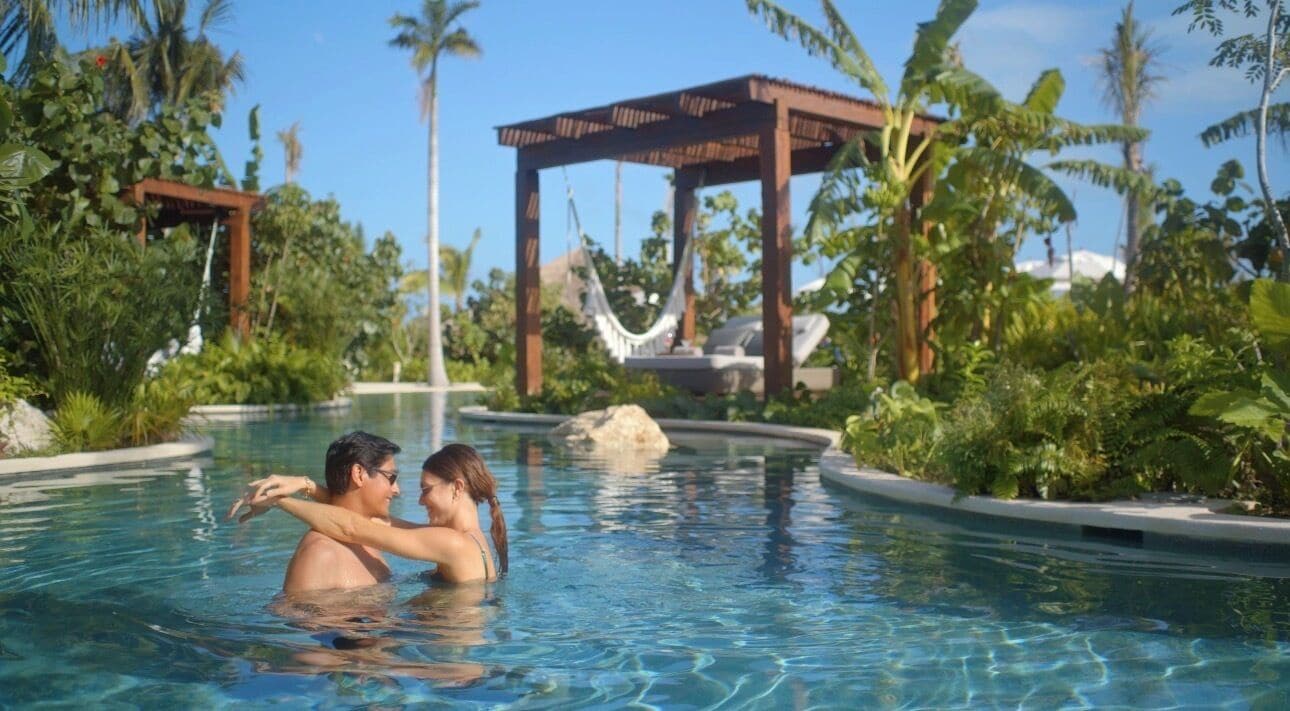 A couple in a clear pool with a wooden gazebo and lounge chairs in the background.
