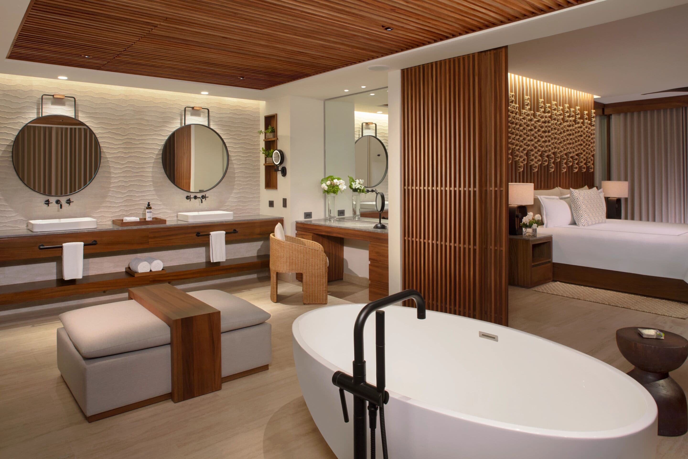 A modern hotel room with a large oval bathtub, a double vanity with round mirrors, a wooden ceiling, and a large bed with a headboard.