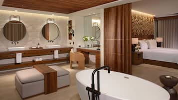 A modern hotel room with a large oval bathtub, a double vanity with round mirrors, a wooden ceiling, and a large bed with a headboard.